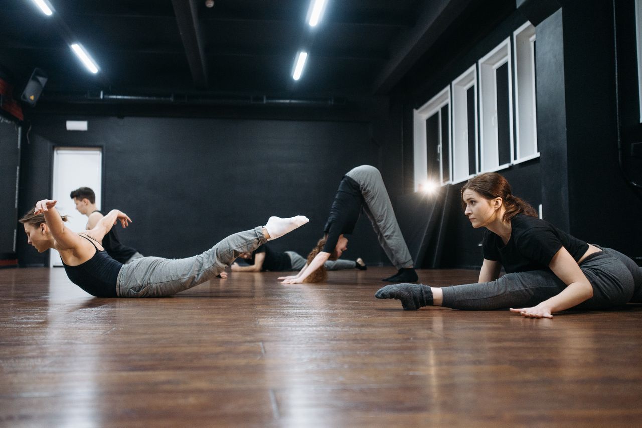 class-02 A group of adults practicing dance flexibility exercises together indoors on a wooden floor.